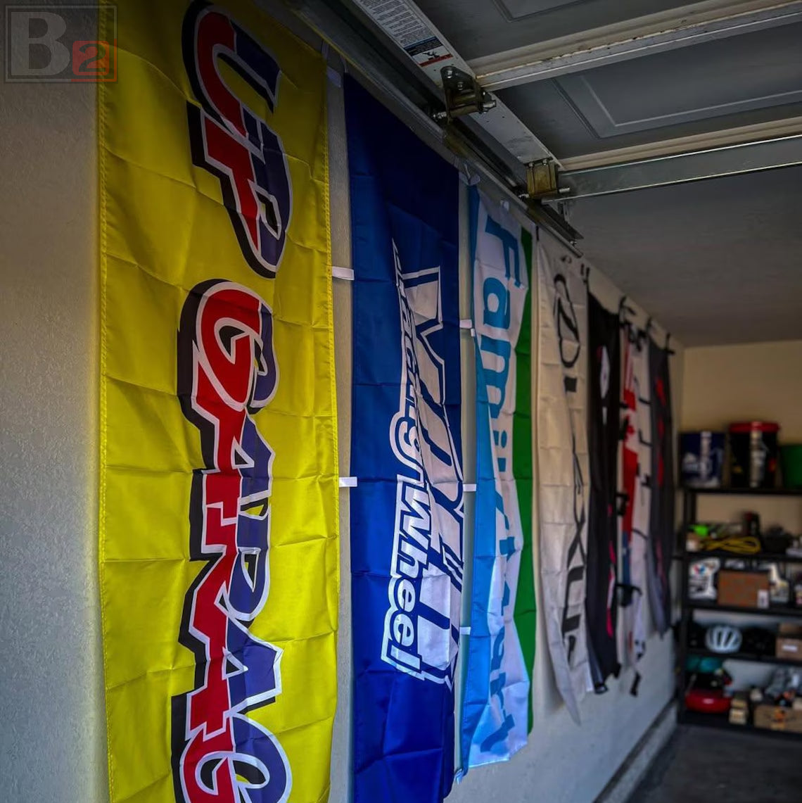 A row of vertical racing flags hanging on a garage door track in a garage. The most prominent flag is bright yellow with 'UP GARAGE' in red and blue lettering, followed by blue and white flags.