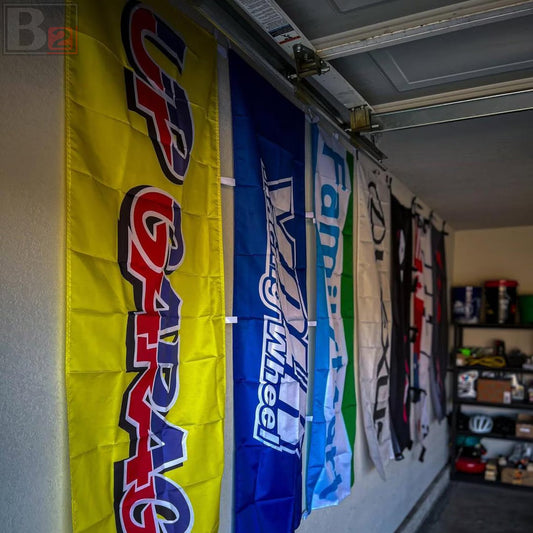 A row of vertical racing flags hanging on a garage door track in a garage. The most prominent flag is bright yellow with 'UP GARAGE' in red and blue lettering, followed by blue and white flags.